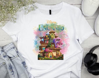 May include: White t-shirt featuring the Disney Encanto logo and a colorful illustration of the Madrigal family's house. The design includes the word "Encanto" in a vibrant, multi-colored font, with a watercolor-style background.