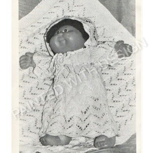 Könnte beinhalten: Ein Schwarzweißfoto einer Cabbage Patch Kid Puppe, die ein gehäkeltes Kleid und eine Häkelmütze trägt. Die Puppe liegt auf einer gehäkelten Decke.