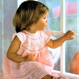 May include: A little girl wearing a pink crocheted dress with a scalloped neckline and a matching pair of crocheted shoes.