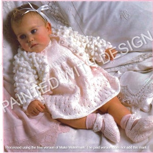 以下が含まれることがあります： ボブルステッチの模様が施された、白のニット製ベビーカーディガン。カーディガンは、ピンクのニットドレスとそれに合わせたブーティを履いた赤ちゃんが着ています。
