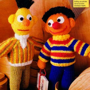 May include: Two hand-knitted dolls representing Bert and Ernie. The yellow doll wears a striped jumper and olive trousers. The orange doll has black hair and wears a blue, yellow, and red striped jumper with blue trousers, holding a small book.