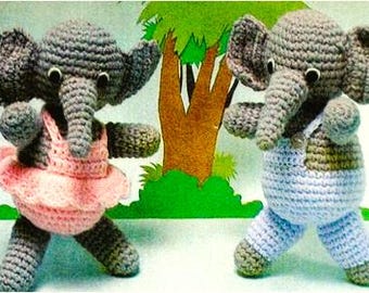 May include: Two grey crocheted elephant dolls. The left elephant wears a pink crocheted tutu and top. The right elephant wears light blue crocheted overalls with straps. Both dolls have black and white plastic eyes and trunks.