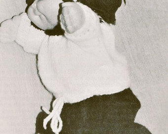 May include: A doll with a round face and dark yarn hair wearing a white knit sweater and dark pants with a white drawstring waist. The pants feature two white horizontal stripes near the cuffs. The doll is captured in a black and white image.