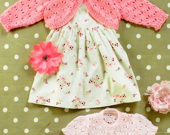 May include: A white sleeveless dress with a small red and pink floral print, paired with a bright pink knit bolero, a pale pink knit bolero, a large pink fabric flower, and a pair of light pink shoes with a scalloped edge and a single strap.