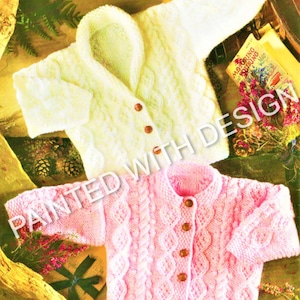 Puede incluir: Dos cárdigans de punto para bebé, uno blanco y otro rosa, con un patrón de trenza y botones.