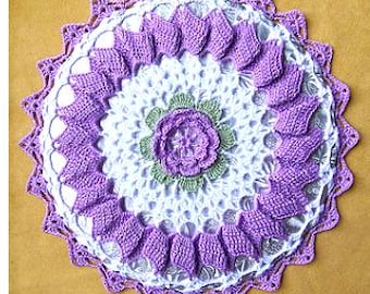 May include: A round, handcrafted crochet doily featuring a white lace base with a central purple flower surrounded by light green leaves. The outer edge is decorated with a ring of textured, raised purple petal-like shapes and a scalloped border.