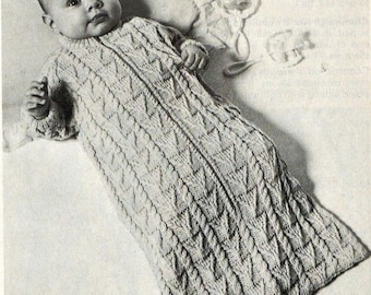 May include: A black and white photograph of a baby wearing a long-sleeved, cable-knit sleeping bag with a central zipper closure. The garment features a textured pattern of vertical cables and diamond shapes, with ribbed cuffs at the wrists.