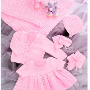 May include: A set of light pink knitted baby items including a textured blanket, a long-sleeved cardigan with buttons, a short-sleeved dress, a bonnet, and a pair of booties. A small gray plush elephant toy is placed on the blanket.