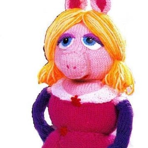May include: A hand-knitted doll featuring a pig-like character with bright yellow yarn hair, blue eyes, and long purple sleeves. The figure wears a vibrant pink dress with a white collar and small red accents, crafted with detailed yarn textures.