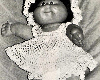 May include: A black and white photograph of a doll with a round face, dark hair, and dimpled cheeks. The doll wears a white crocheted bonnet with a ruffled edge and a matching white crocheted dress with a flared skirt and intricate pattern.
