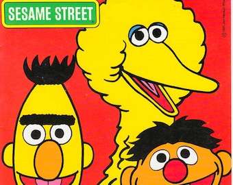May include: A vibrant red graphic print featuring the Sesame Street logo in a green rectangular sign, alongside illustrations of Big Bird, Bert, and Ernie. The characters are depicted with their signature yellow, orange, and black features.