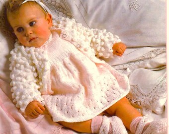 May include: A pale pink knitted dress with a matching white bobble-stitch cardigan, white knitted booties, and a white ribbon hair accessory. The outfit features a textured pattern on the cardigan and a delicate knit design on the dress.