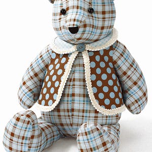 May include: A stuffed teddy bear made of light blue and brown plaid fabric. The bear wears a brown vest with light blue polka dots, trimmed with cream-colored lace and a single blue button at the collar. It has black bead eyes and a black nose.