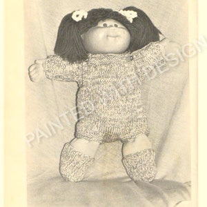 May include: A black and white photo of a Cabbage Patch Kid doll wearing a knitted sweater and pants. The doll has a knitted hat with white bows on top.