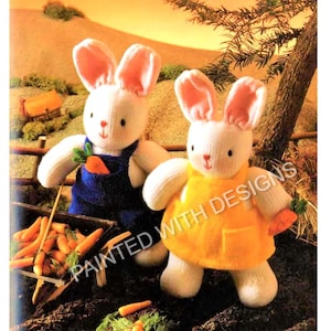 May include: Two white knitted bunny rabbits with pink ears. One bunny is wearing blue overalls and holding a carrot. The other bunny is wearing a yellow dress and holding a carrot. Both bunnies are sitting in a garden setting with carrots in a wheelbarrow.