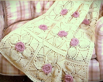 May include: A cream-colored crochet blanket featuring a repeating square pattern with intricate openwork details. Each square is adorned with a small, three-dimensional dusty pink crocheted flower attached to the center of the textured fabric.