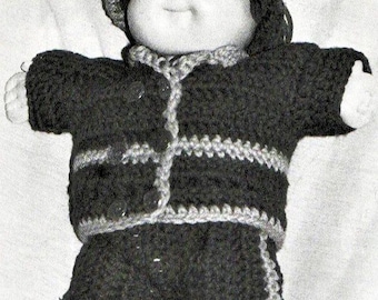 May include: A doll with a round face and dark hair wearing a crocheted outfit consisting of a dark jacket with light-colored trim, matching dark pants with light-colored side stripes, a matching dark hat, and light-colored crocheted shoes.