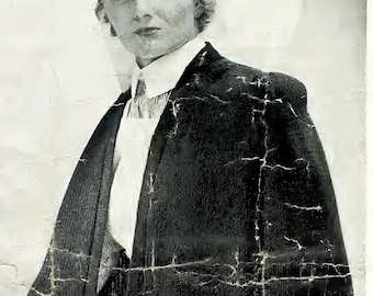 May include: A vintage black and white photograph showing a person wearing a dark, textured cape over a collared shirt and a light-colored apron. The person has a short, wavy hairstyle and is wearing a head covering, captured in a portrait style.