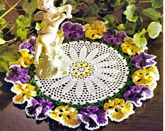 May include: A round white crocheted doily featuring a central sunburst pattern and a decorative border of alternating purple and yellow pansy flowers. A small white cherub figurine stands on the left side of the circular lace mat.
