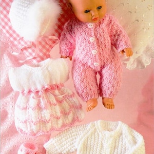 May include: A baby doll wearing a pink knitted long-sleeve romper with white buttons. The set includes a white knitted dress with pink ribbon trim, a white knitted cardigan, a white bonnet with faux fur trim, and two white knitted booties with pink ribbons.