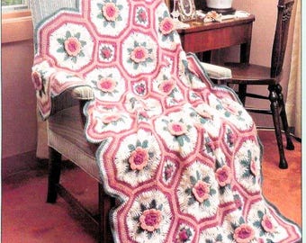 May include: A crocheted blanket featuring a repeating pattern of white octagonal motifs with dark green leaf accents and raised, three-dimensional pink floral centers. The blanket is bordered by rows of pink and dark green yarn in a scalloped edge.