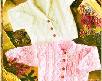 May include: Two knitted cardigans, one white and one pink, featuring intricate cable knit patterns and textured stitch details. The white cardigan has three brown buttons, while the pink cardigan features five brown buttons down the front.