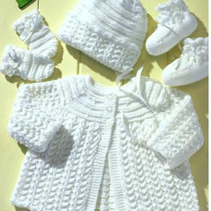 Peut inclure: Un ensemble tricoté blanc pour bébé comprenant un gilet à manches longues avec fermeture à boutons, un bonnet assorti, une paire de chaussons avec rubans et une paire de moufles avec rubans, le tout avec un motif en maille torsadée.