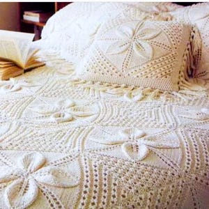 May include: A cream-colored crocheted bedspread and a matching square throw pillow featuring a repeating floral motif. The textile design consists of intricate knit patterns with raised petal shapes arranged in a grid, finished with fringe.