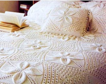 May include: A cream-colored crocheted bedspread and a matching square throw pillow featuring a repeating floral motif. The textile design consists of intricate knit patterns with raised petal shapes arranged in a grid, finished with fringe.