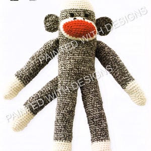 Puede incluir: Un mono de calcetín tejido a mano con boca roja, cuerpo marrón y blanco, y detalles crema. El mono tiene brazos y piernas largos, y el texto "Basic Crochet Sock Monkey" es visible.