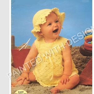 May include: A baby girl wearing a yellow crocheted dress and matching sun hat. The baby is sitting on a sandy beach.