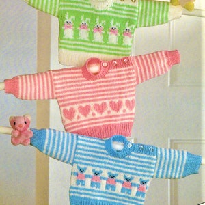 May include: Three knitted baby sweaters in different colours and patterns. The first sweater is green and white with a bunny pattern. The second sweater is pink and white with a heart pattern. The third sweater is blue and white with a bear pattern.