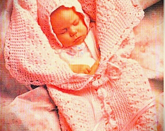 May include: A light pink crocheted baby bunting with a matching hood and decorative ribbon ties. The soft, textured knit fabric wraps around a sleeping infant, featuring a delicate scalloped edge and a central vertical row of small pom-poms.