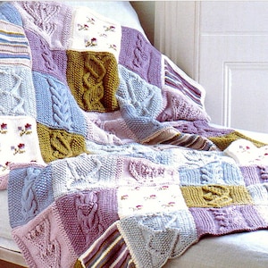 Può includere: Una coperta in maglia patchwork composta da pannelli quadrati nei toni del lavanda, azzurro, verde oliva e bianco. Il design presenta diverse texture come motivi a treccia, punto riso e quadrati bianchi con piccoli motivi floreali.