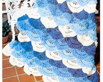 May include: A crochet blanket featuring a repeating fan-shaped pattern in shades of cream, light blue, and dark blue. The design consists of textured, scalloped rows with openwork detailing, creating a layered, dimensional appearance throughout.