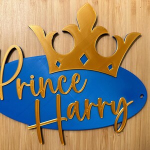 May include: A blue oval sign with the words "Prince Harry" in gold script, topped with a gold crown. The sign has two small holes for hanging. The background is a light brown wood.