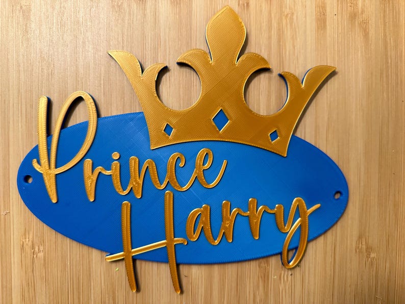 May include: A blue oval sign with the words "Prince Harry" in gold script, topped with a gold crown. The sign has two small holes for hanging. The background is a light brown wood.