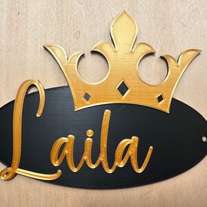 May include: A black oval sign with the name "Laila" in gold script, topped with a gold crown. The sign has two holes for hanging and is set against a light wood background.