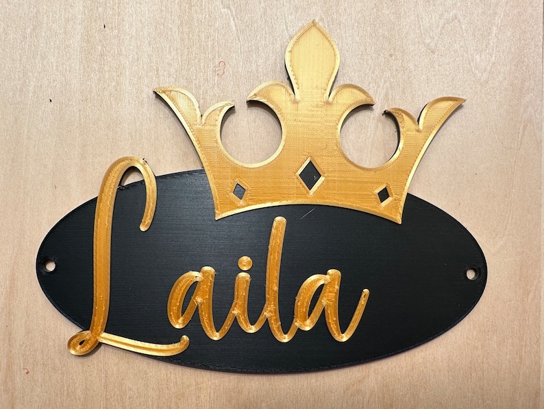 May include: A black oval sign with the name "Laila" in gold script, topped with a gold crown. The sign has two holes for hanging and is set against a light wood background.