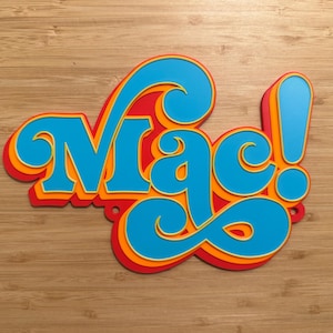 Puede incluir: Un letrero en capas con la palabra "Mac!" en una fuente retro. Las letras son azules con contornos naranjas y rojos, creando un efecto 3D. El signo de exclamación también es azul. El letrero está sobre una superficie de madera.