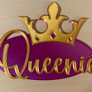 May include: A decorative sign featuring the name "Queenie" in gold script on a purple oval, topped with a gold crown. The sign is designed with a 3D printed appearance and includes two small holes for hanging.