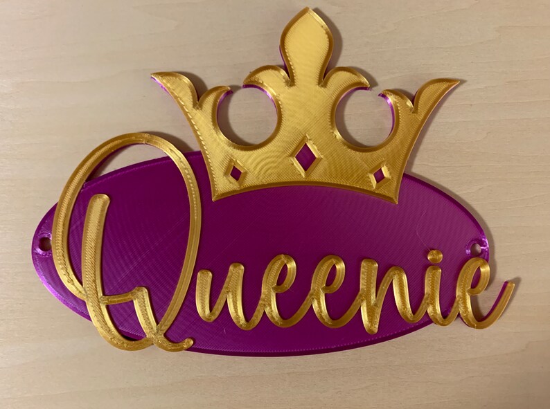 May include: A decorative sign featuring the name "Queenie" in gold script on a purple oval, topped with a gold crown. The sign is designed with a 3D printed appearance and includes two small holes for hanging.
