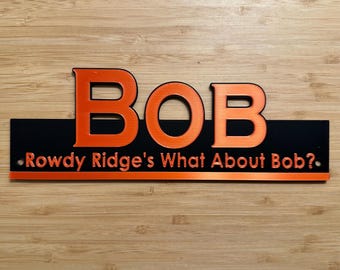 May include: A rectangular black sign with orange lettering featuring the large text BOB at the top and the smaller text Rowdy Ridge's What About Bob? below. The sign has a horizontal orange stripe at the bottom and two small mounting holes.