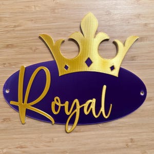 May include: A gold-colored crown sits atop a purple oval with the word "Royal" in gold script. The design has a regal theme, suitable for decorations or signage. The background is a light brown wooden surface.