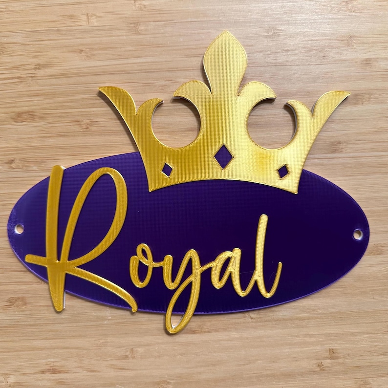 May include: A gold-colored crown sits atop a purple oval with the word "Royal" in gold script. The design has a regal theme, suitable for decorations or signage. The background is a light brown wooden surface.