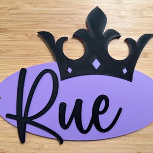 May include: A black crown sits atop a lavender oval with the name "Rue" in black cursive. The crown has a decorative design. The background is a light brown wooden surface.
