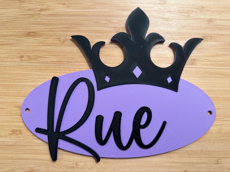 May include: A black crown sits atop a lavender oval with the name "Rue" in black cursive. The crown has a decorative design. The background is a light brown wooden surface.