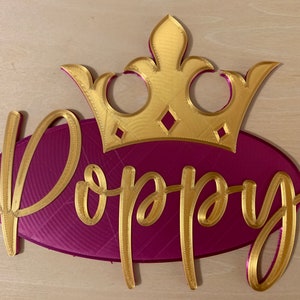 May include: A gold-colored crown sits atop a magenta oval with the name "Poppy" in gold script. The crown has diamond-shaped accents. This decorative piece is likely a personalized name sign.