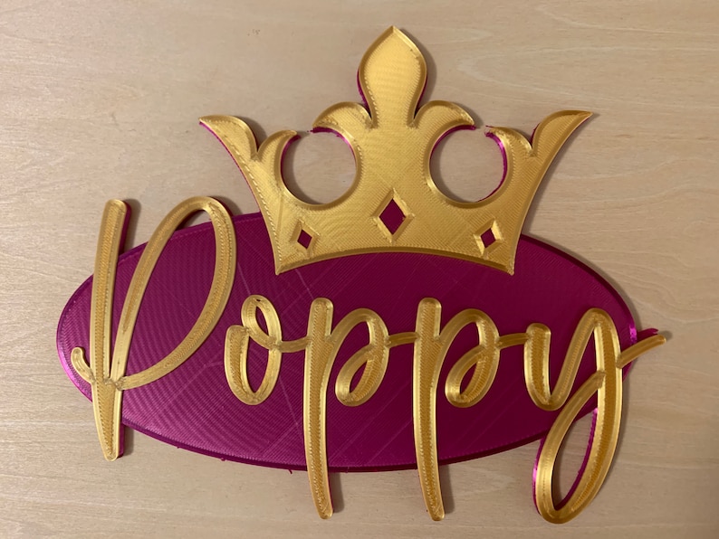 May include: A gold-colored crown sits atop a magenta oval with the name "Poppy" in gold script. The crown has diamond-shaped accents. This decorative piece is likely a personalized name sign.