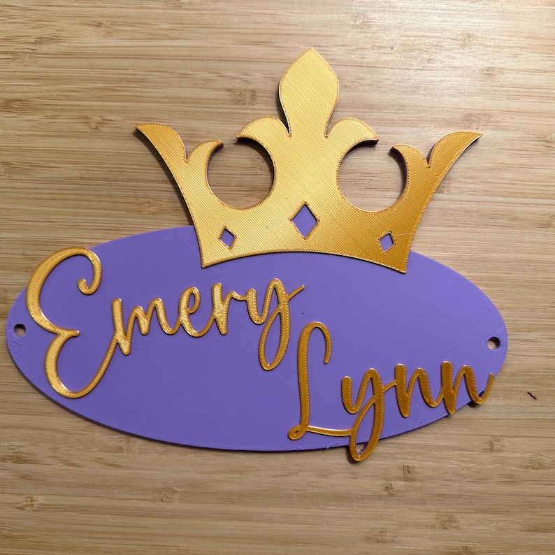 May include: A gold-colored crown sits above a purple oval sign. The sign features the name "Emery Lynn" in gold script. The sign has two small holes, likely for hanging. The background is a light brown wood grain.